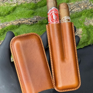 May include: A brown leather cigar case, open to reveal two cigars with colorful labels. The case is a rich, medium brown with visible stitching around the edges. The background is a blurred mix of green and black.