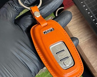 Könnte beinhalten: Orangener Leder-Autoschlüsselanhänger mit silbernem Schlüsselring. Der Schlüsselanhänger hat silberne Knöpfe und eine genähte Bordüre. Der Schlüsselanhänger wird von einer behandschuhten Hand gehalten.