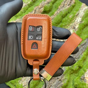 May include: A tan leather car key fob cover with a matching strap and silver key ring. The cover has embossed icons for lock, unlock, lights, and boot release. The fob is held by a black gloved hand.