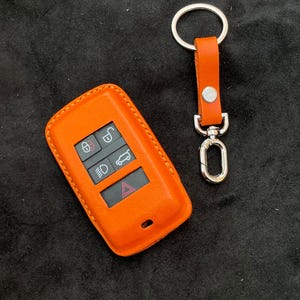 May include: Orange leather car key fob cover and matching key ring. The key fob cover has buttons for locking, unlocking, lights, and a trunk release. The key ring has a silver clasp and a leather strap with a silver rivet.