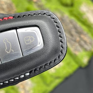 Op de afbeelding: Close-up van een zwarte leren sleutelhanger met zilveren knoppen. De sleutelhanger heeft rode stiksels aan de bovenkant en witte stiksels langs de randen. De knoppen hebben auto- en slotpictogrammen.