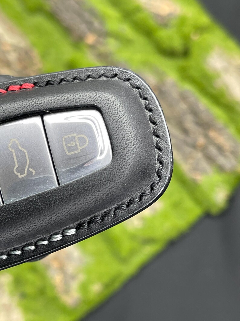 Op de afbeelding: Close-up van een zwarte leren sleutelhanger met zilveren knoppen. De sleutelhanger heeft rode stiksels aan de bovenkant en witte stiksels langs de randen. De knoppen hebben auto- en slotpictogrammen.