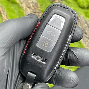 Op de afbeelding: Zwarte leren autosleutelhoes met rode en witte stiksels. De sleutelhanger heeft drie zilveren knoppen en een leren riem met een metalen sluiting. De achtergrond is een wazig beeld van groen gebladerte.
