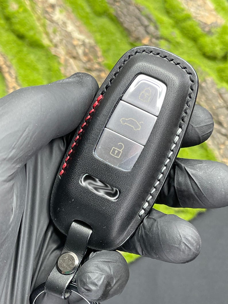 Op de afbeelding: Zwarte leren autosleutelhoes met rode en witte stiksels. De sleutelhanger heeft drie zilveren knoppen en een leren riem met een metalen sluiting. De achtergrond is een wazig beeld van groen gebladerte.