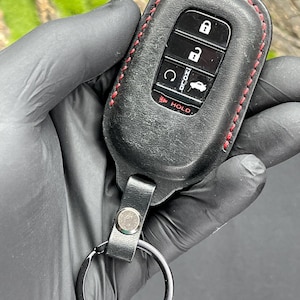 May include: Black leather key fob cover with red stitching. The cover has a key ring and a button to hold the key fob. The key fob has buttons for lock, unlock, and other functions. The word "HOLD" is printed on the key fob.