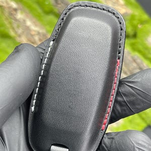 Op de afbeelding: Zwarte leren sleuteletui met witte en rode stiksels. De etui heeft een rechthoekige vorm met afgeronde randen en een klein bandje aan de onderkant. De stiksels zijn zichtbaar langs de randen van de etui.