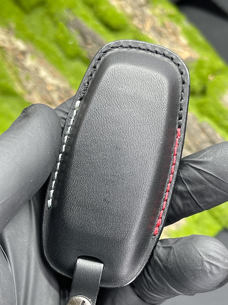 Op de afbeelding: Zwarte leren sleuteletui met witte en rode stiksels. De etui heeft een rechthoekige vorm met afgeronde randen en een klein bandje aan de onderkant. De stiksels zijn zichtbaar langs de randen van de etui.