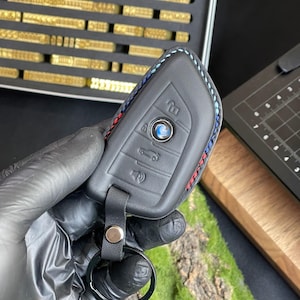 May include: Black leather car key fob cover with blue, red, and white stitching. The fob has buttons for locking, unlocking, and other car functions. A black key ring is attached to the fob.