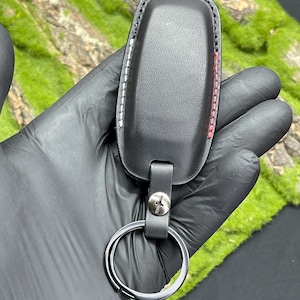 Op de afbeelding: Zwarte leren sleutelhangerhoes met witte en rode stiksels. De sleutelhanger heeft een zilveren sleutelring bevestigd met een leren bandje en een zilveren schroef. De achtergrond is mosgroen.