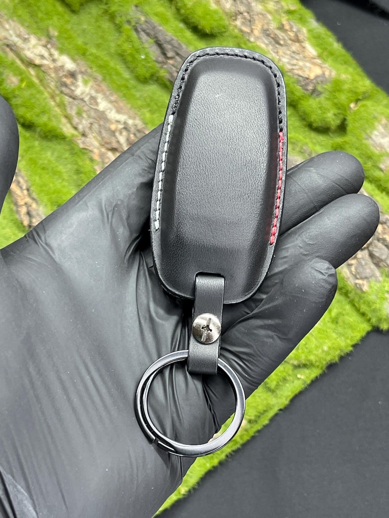 Op de afbeelding: Zwarte leren sleutelhangerhoes met witte en rode stiksels. De sleutelhanger heeft een zilveren sleutelring bevestigd met een leren bandje en een zilveren schroef. De achtergrond is mosgroen.