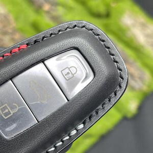 Op de afbeelding: Zwarte leren sleutelhangerhoes met rode en witte stiksels. De sleutelhanger heeft drie zilveren knoppen met pictogrammen voor het vergrendelen, ontgrendelen en openen van de auto. De hoes is ontworpen om de autosleutel te beschermen.