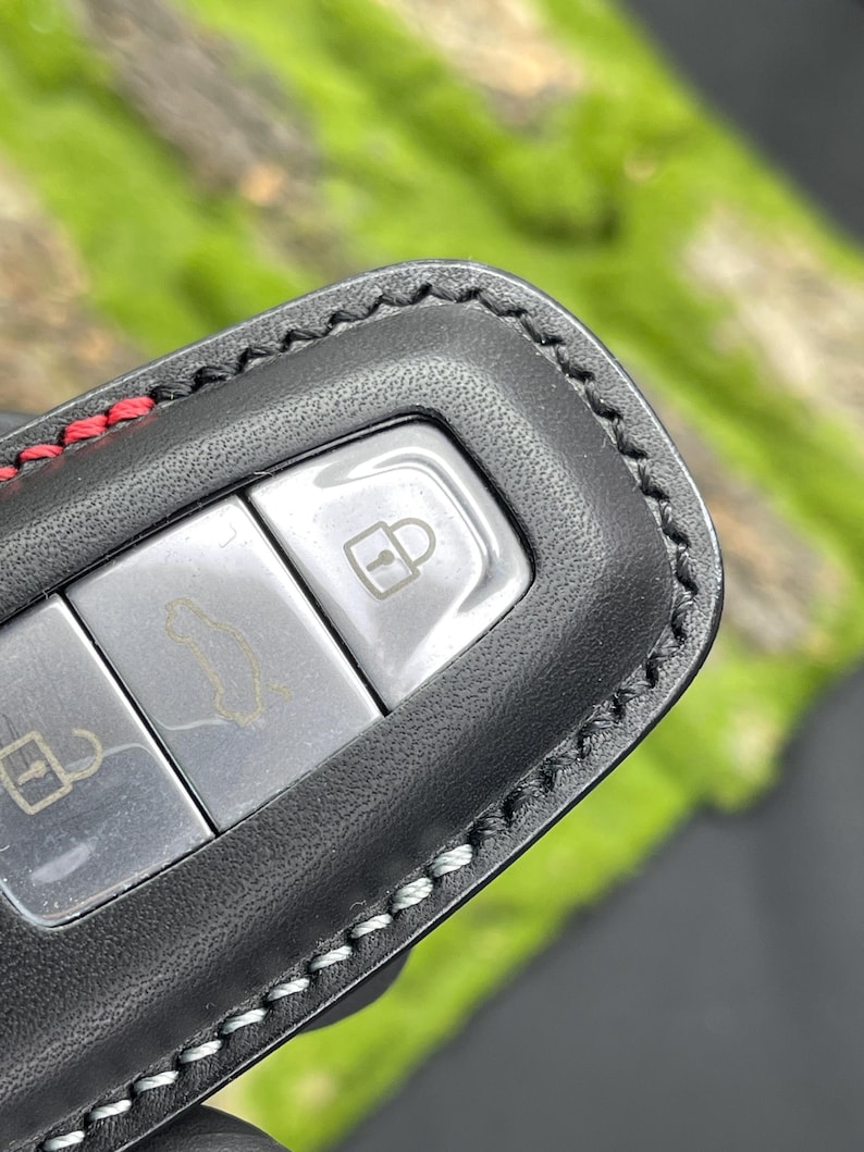 Op de afbeelding: Zwarte leren sleutelhangerhoes met rode en witte stiksels. De sleutelhanger heeft drie zilveren knoppen met pictogrammen voor het vergrendelen, ontgrendelen en openen van de auto. De hoes is ontworpen om de autosleutel te beschermen.