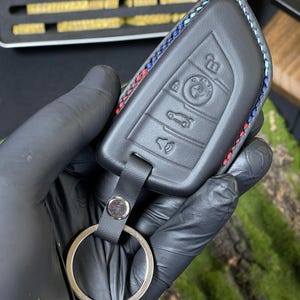 May include: Black leather BMW key fob cover with red, blue, and white stitching. The fob has embossed buttons and a metal keyring attachment. The cover is held by a gloved hand.
