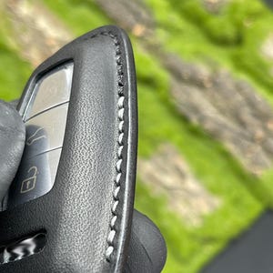 Op de afbeelding: Close-up van een zwarte leren sleutelhangerhoes met witte stiksels. De sleutelhanger heeft drie knoppen en een sleutelsymbool. De achtergrond is wazig groen en bruin.