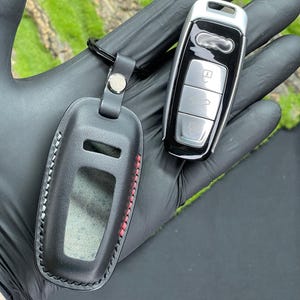Op de afbeelding: Zwarte leren sleutelhangerhoes met rode stiksels en een zilveren drukknoopsluiting, naast een zwarte en zilveren autosleutel. De sleutelhangerhoes heeft een doorzichtig venster. De autosleutel heeft drie zilveren knoppen.