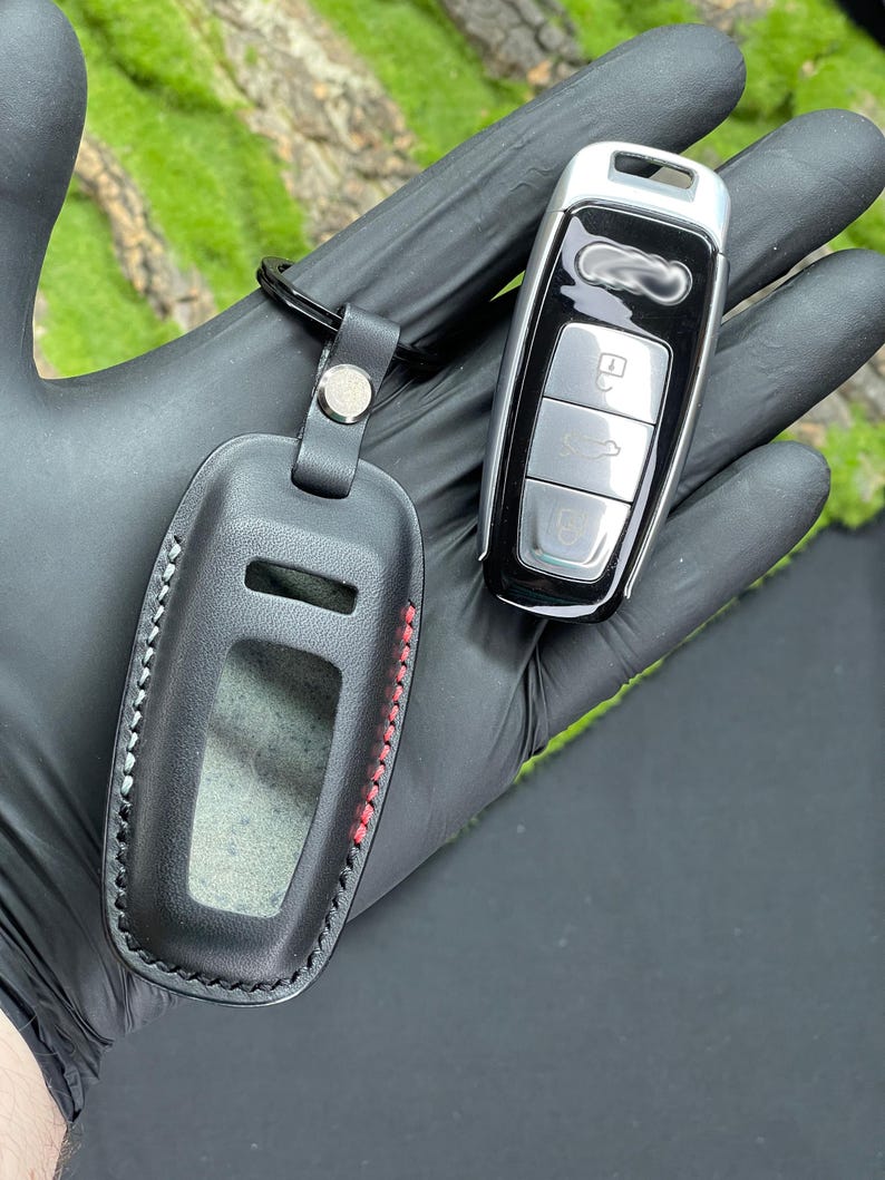 Op de afbeelding: Zwarte leren sleutelhangerhoes met rode stiksels en een zilveren drukknoopsluiting, naast een zwarte en zilveren autosleutel. De sleutelhangerhoes heeft een doorzichtig venster. De autosleutel heeft drie zilveren knoppen.