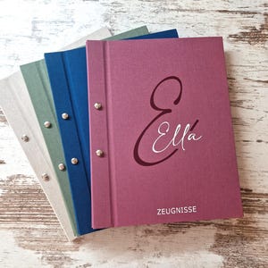 Könnte beinhalten: Ein Stapel aus vier stoffbezogenen Mappen mit Metallschrauben. Die oberste Mappe ist dunkelrosa und mit dem Namen Ella sowie dem Wort ZEUGNISSE in Weiß bedruckt. Die weiteren Mappen sind in Blau, Salbeigrün und Hellgrau gehalten und haben Metallnieten.