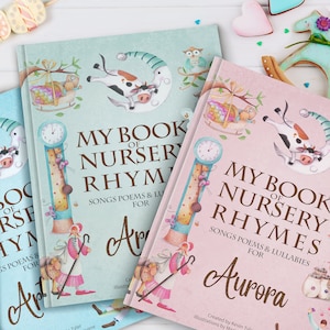 May include: Two personalised children's books with pastel blue and pink covers. The covers feature illustrations of a cow, a clock, and a bird. The text on the covers reads "My Book of Nursery Rhymes" and includes the names "Aria" and "Aurora".