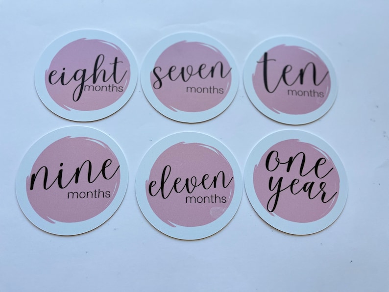 May include: Six pink and white circular baby milestone stickers with black text. The stickers say "eight months", "seven months", "ten months", "nine months", "eleven months", and "one year".
