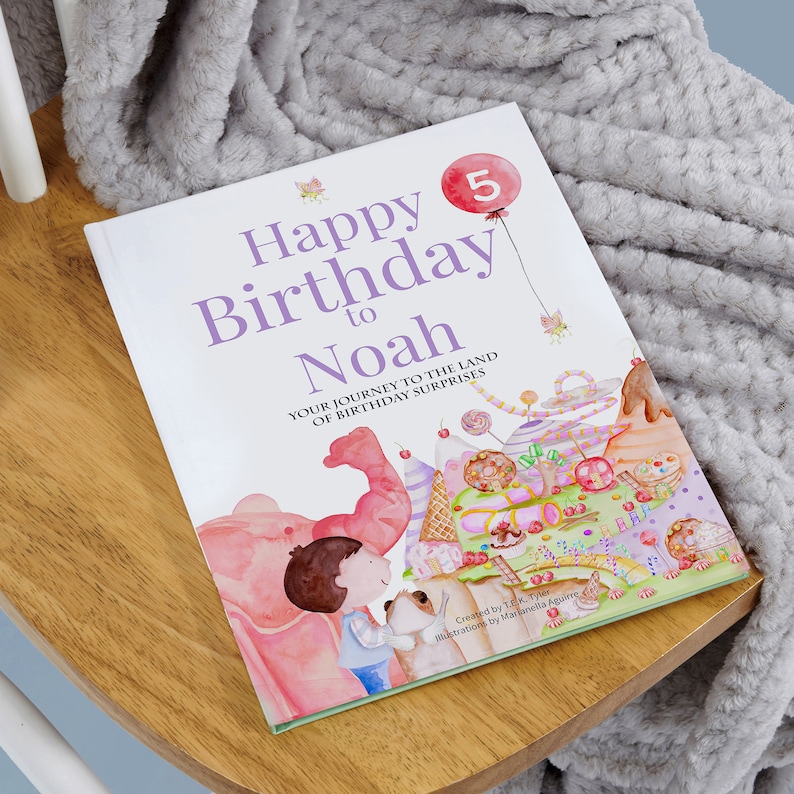 Pode incluir: Um livro branco com uma ilustra&ccedil;&atilde;o colorida de um bolo de anivers&aacute;rio e um elefante rosa. A capa do livro diz "Happy Birthday to Noah" e "Your Journey to the Land of Birthday Surprises".