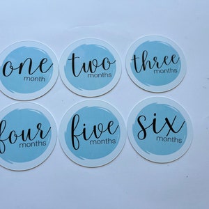 May include: Six blue and white baby milestone stickers with the numbers one through six written in black cursive. Each sticker has the word "months" written below the number.