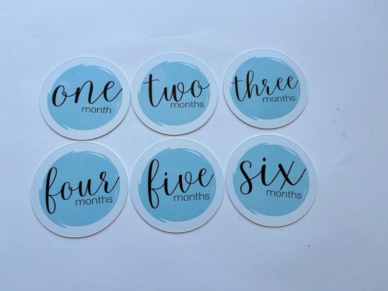 May include: Six blue and white baby milestone stickers with the numbers one through six written in black cursive. Each sticker has the word "months" written below the number.