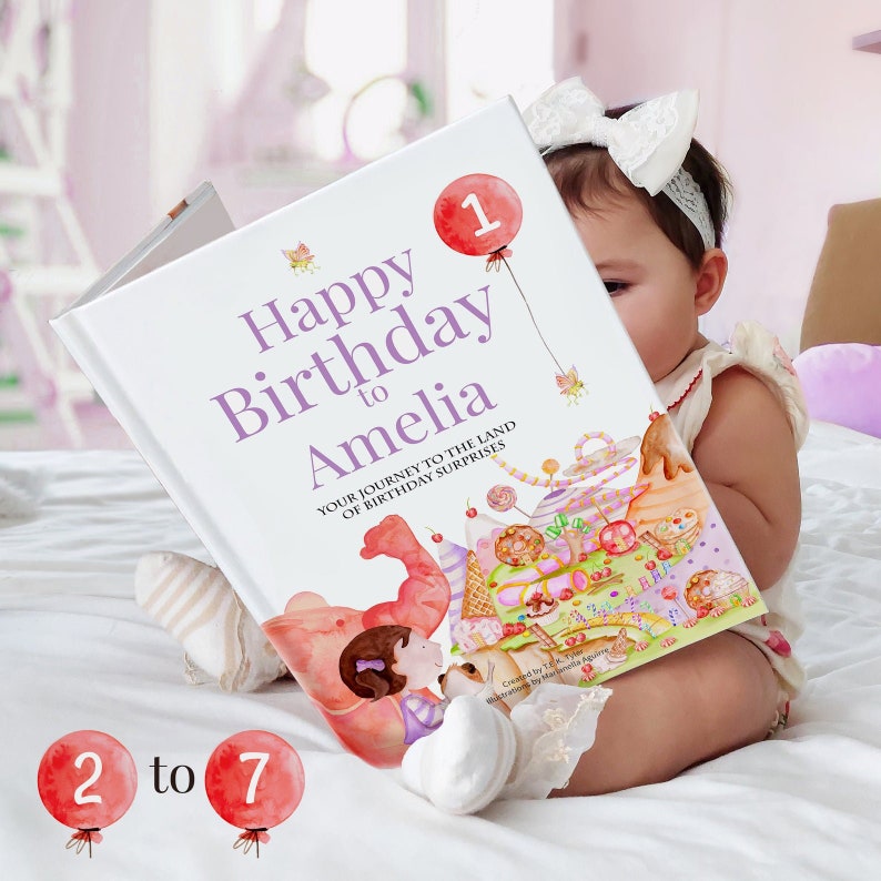 Pode incluir: Um livro branco com as palavras "Happy Birthday to Amelia" e uma ilustra&ccedil;&atilde;o colorida de um bolo de anivers&aacute;rio e bal&otilde;es. O livro est&aacute; sendo segurado por um beb&ecirc; que est&aacute; usando uma faixa de cabe&ccedil;a branca e um vestido branco.