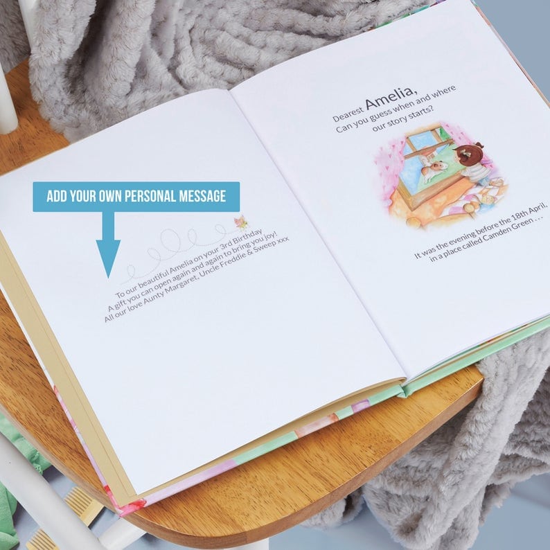 Pode incluir: Um livro infantil personalizado com um design floral rosa e verde. O livro est&aacute; aberto em uma p&aacute;gina com uma mensagem escrita &agrave; m&atilde;o que diz "To our beautiful Amelia on your 3rd Birthday A gift you can open again and again to bring you joy! All our love Aunty Margaret, Uncle Freddie & Sweep xxx". A p&aacute;gina seguinte tem uma ilustra&ccedil;&atilde;o de uma menina olhando para fora de uma janela e o texto "Dearest Amelia, Can you guess when and where our story starts? It was the evening before the 18th April, in a place called Camden Green..."