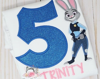 Puede incluir: Una camiseta blanca con un número cinco grande de color azul brillante, un personaje de conejo de dibujos animados bordado con uniforme de policía y el nombre Trinity bordado en letras rosas, con un pequeño logotipo de Sew Younique Handmades.