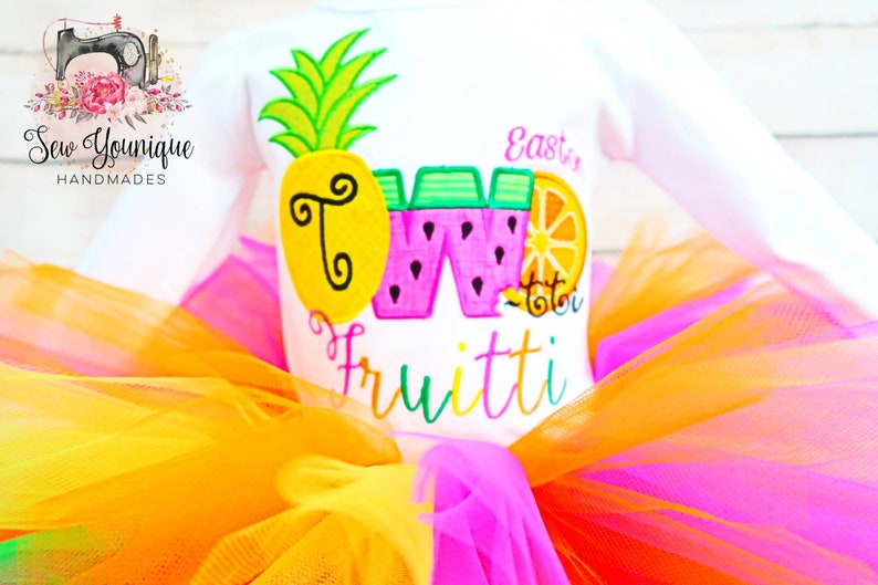 Puede incluir: Un body blanco con un dise&ntilde;o bordado de colores de una pi&ntilde;a, una sand&iacute;a y una naranja. El body dice "Eastin Two Frutti" en una fuente de colores. El body se combina con un tut&uacute; de tul multicolor.