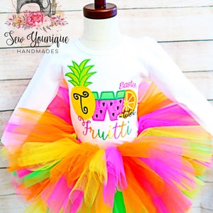 Puede incluir: Una camiseta blanca de manga larga con un dise&ntilde;o de pi&ntilde;a, sand&iacute;a y naranja. La camiseta dice "Two Fruitti" y "Easton" en letras rosas. La camiseta se combina con un tut&uacute; de tul multicolor.