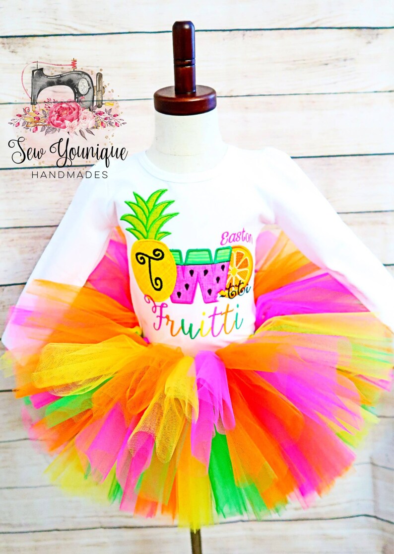 Puede incluir: Una camiseta blanca de manga larga con un dise&ntilde;o de pi&ntilde;a, sand&iacute;a y naranja. La camiseta dice "Two Fruitti" y "Easton" en letras rosas. La camiseta se combina con un tut&uacute; de tul multicolor.