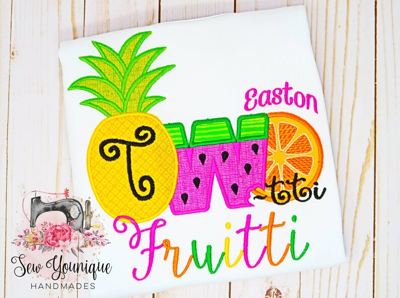 Puede incluir: Una camiseta blanca con un dise&ntilde;o bordado de colores que incluye una pi&ntilde;a, una sand&iacute;a y una naranja. El dise&ntilde;o dice "Two Fruitti" y "Easton" en letras rosas.