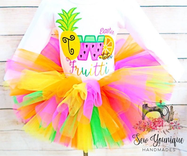 Puede incluir: Una camisa blanca de manga larga con un dise&ntilde;o bordado de colores de una pi&ntilde;a, una sand&iacute;a y un lim&oacute;n. La camisa dice "Easton" y "Dos" con un dise&ntilde;o de fruta. La camisa se combina con un tut&uacute; de tul multicolor.