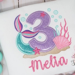 May include: White t-shirt with a mermaid-themed design. Features a purple glittery number 3, a mermaid tail, seashells, and the name "Melia" in pink script. Includes bubbles and coral accents. The design is embroidered.