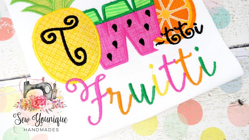 Puede incluir: Una camisa blanca con dise&ntilde;os de frutas bordadas de colores. La camisa dice "So Fruitti" en una fuente de letra cursiva de colores.