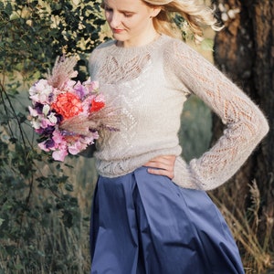 May include: A woman wearing a light gray knitted sweater with a delicate lace pattern and a blue skirt holds a bouquet of pink, purple, and red flowers.