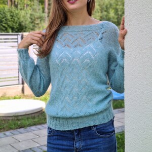 Puede incluir: Un suéter azul claro con cuello barco y mangas largas. El suéter presenta un delicado diseño calado en la parte superior del pecho y un patrón texturizado en el cuerpo. La persona lleva vaqueros azules.