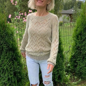 May include: A woman wearing a light green knitted sweater with a delicate lace pattern and distressed light blue jeans. She is standing in a garden setting with green foliage and a fence in the background.