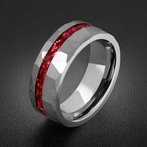 Può includere: Un anello color argento caratterizzato da una superficie esterna martellata e sfaccettata con un intarsio centrale di materiale rosso vivo frantumato che corre orizzontalmente lungo l'intera circonferenza, creando un contrasto di texture e colore.
