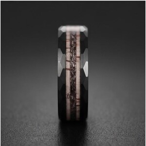 Pu&ograve; includere: Un anello nero con una superficie esterna sfaccettata e un inserto centrale caratterizzato da un materiale testurizzato color marrone chiaro, delimitato da sottili linee marrone scuro. La fascia ha un aspetto metallico scuro e geometrico.