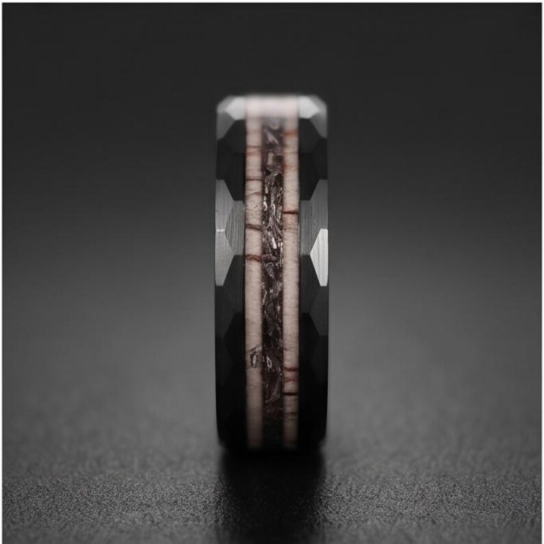 Pu&ograve; includere: Un anello nero con una superficie esterna sfaccettata e un inserto centrale caratterizzato da un materiale testurizzato color marrone chiaro, delimitato da sottili linee marrone scuro. La fascia ha un aspetto metallico scuro e geometrico.