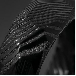 May include: A black object featuring a series of stacked, curved layers with a glossy finish on the top edges and a textured, matte, granular surface on the recessed sections, creating a distinct chevron-like pattern across the structure.