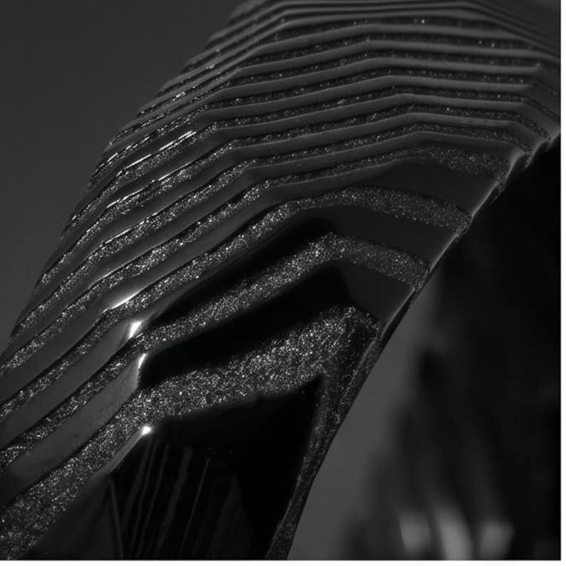 May include: A black object featuring a series of stacked, curved layers with a glossy finish on the top edges and a textured, matte, granular surface on the recessed sections, creating a distinct chevron-like pattern across the structure.