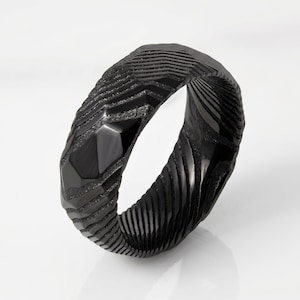 May include: A black ring featuring a textured, multi-faceted surface with alternating matte and polished finishes. The band displays a series of deep, angular grooves and raised ridges that create a complex, geometric pattern around the entire circumference.