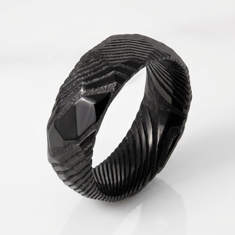 May include: A black ring featuring a textured, multi-faceted surface with alternating matte and polished finishes. The band displays a series of deep, angular grooves and raised ridges that create a complex, geometric pattern around the entire circumference.