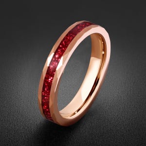 Pode incluir: Um anel em tom de ouro rosa com uma banda exterior facetada e uma incrustação central de material vermelho escuro triturado. O interior da banda é liso e polido, apresentando um acabamento metálico que contrasta com o centro vermelho texturizado.