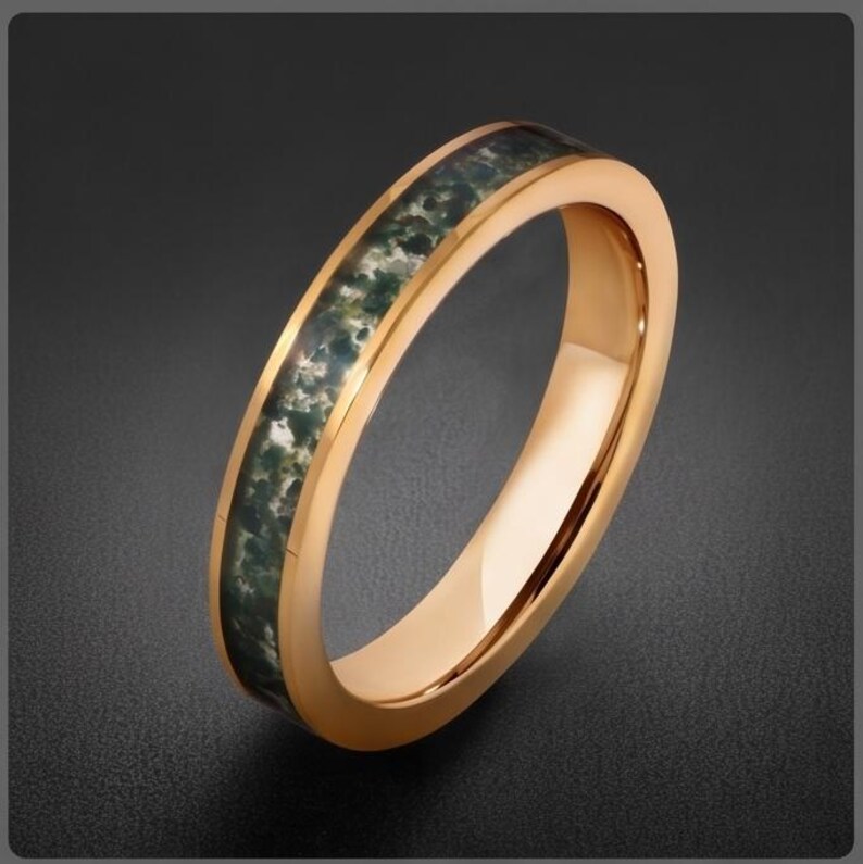 May include: A rose gold-toned band ring featuring a central inlay of dark green and white mottled stone. The metal surface is smooth and polished, contrasting with the textured, organic appearance of the stone inlay running along the center.