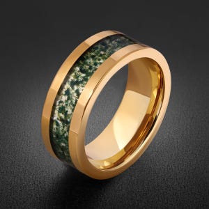 May include: A gold-toned band ring featuring a central inlay of moss agate stone. The polished metal edges frame the textured, dark green and white speckled mineral inlay, creating a smooth, rounded profile with a reflective, metallic finish.