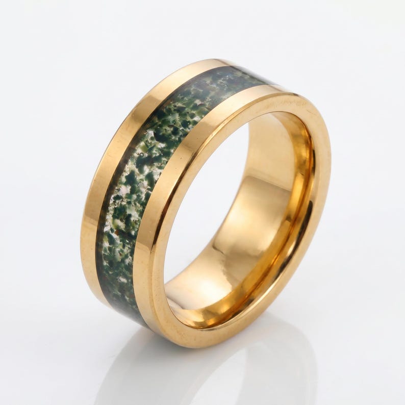 May include: A gold-toned ring featuring a wide, polished band with a central inlay of moss agate. The stone inlay displays a mottled pattern of deep forest green and translucent white inclusions, set within the smooth, reflective metallic surface.